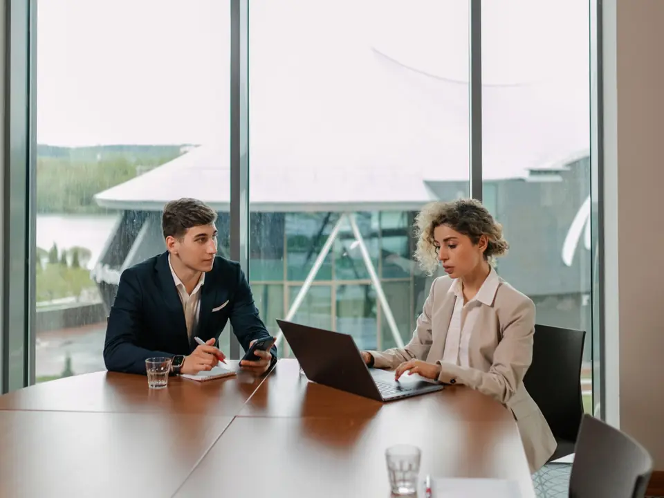 Two people meeting at a conference table with a laptop, in an office with tall windows